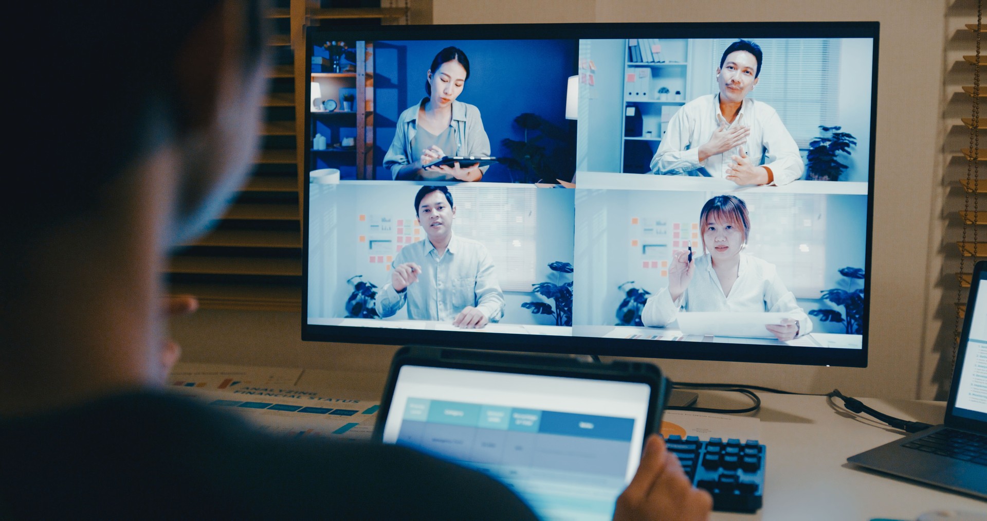Closeup of young Asian man using digital tablet and laptop for video conference with colleagues at night from his home office, showcasing remote work and teamwork. Remotely and finance.